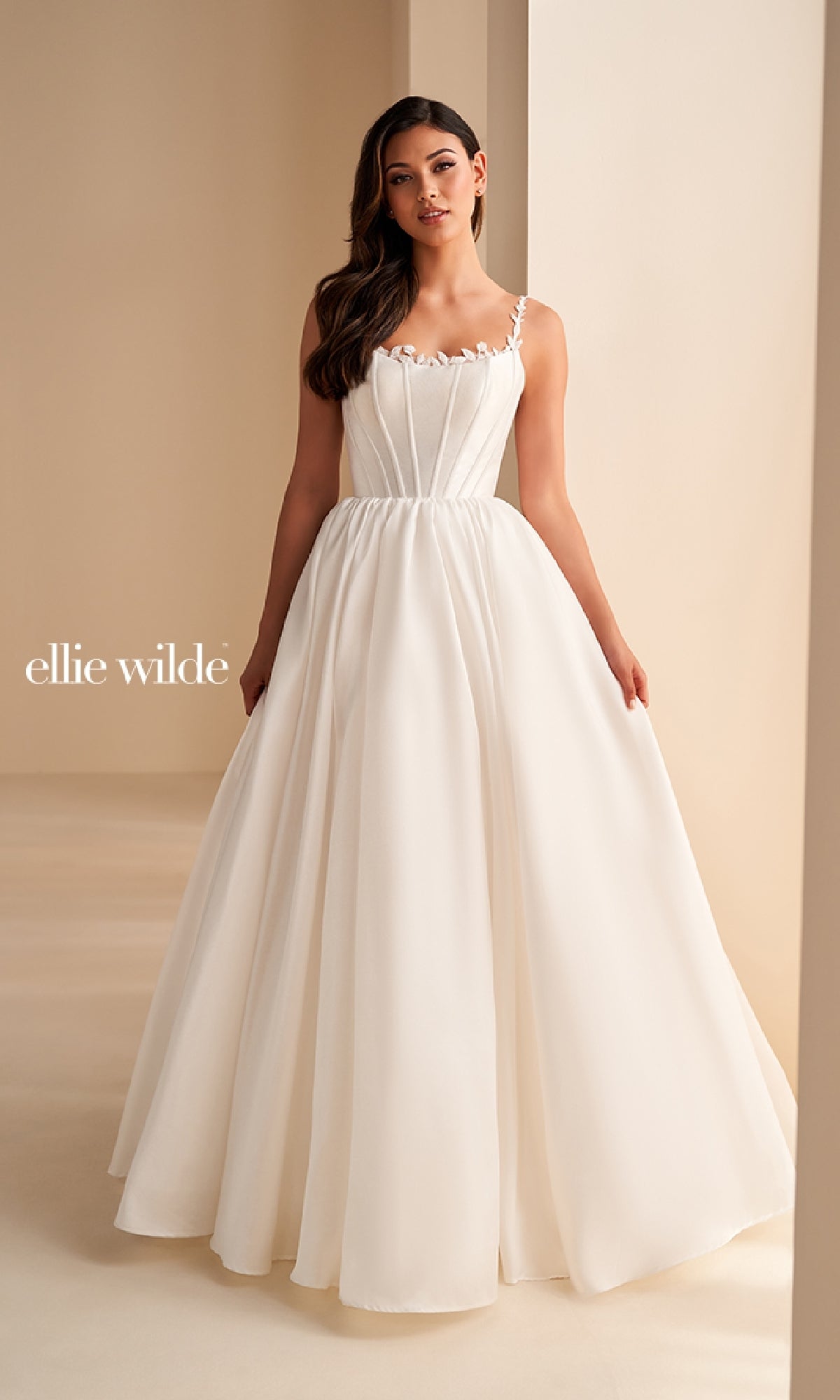 A woman stands indoors wearing the Ellie Wilde EW36221 long prom dress, featuring a white A-line skirt, a fitted sleeveless bodice with delicate straps, subtle pleating, and a simple, elegant design.